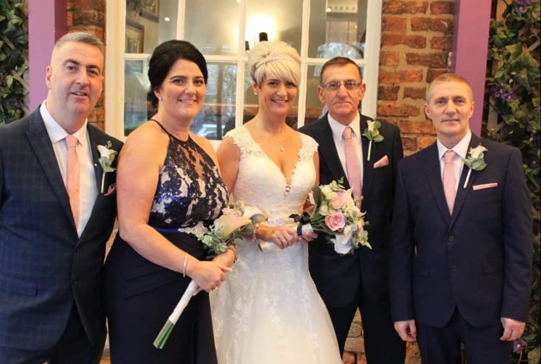 Group of 5 people standing smiling broadly at the camera at a wedding. All are dressed smartly and the bride in a white dress.
