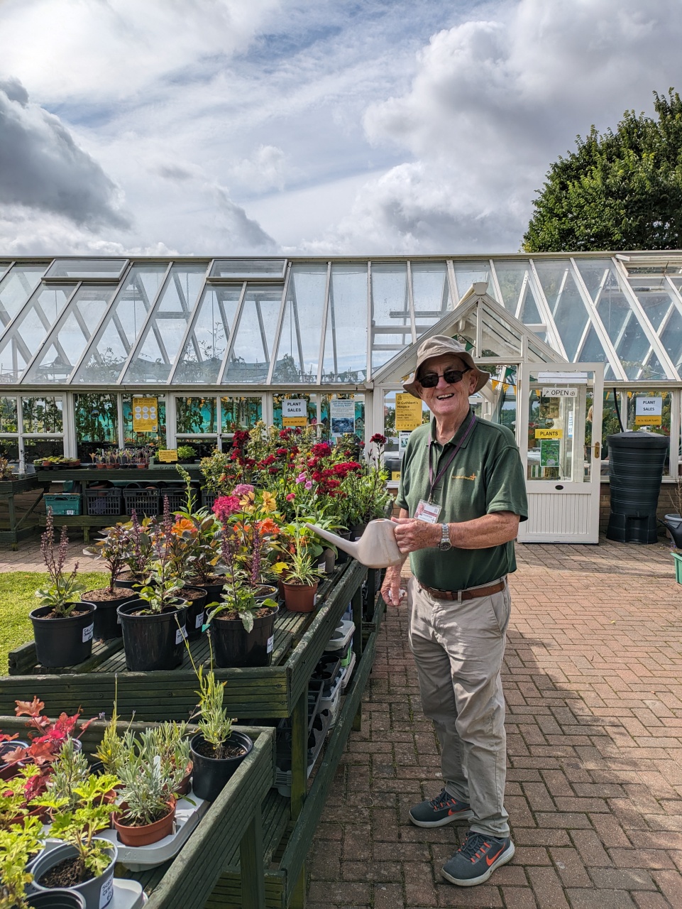 A man stands in front of a greenhouse, watering can in hand, surrounded by plants and sunlight.