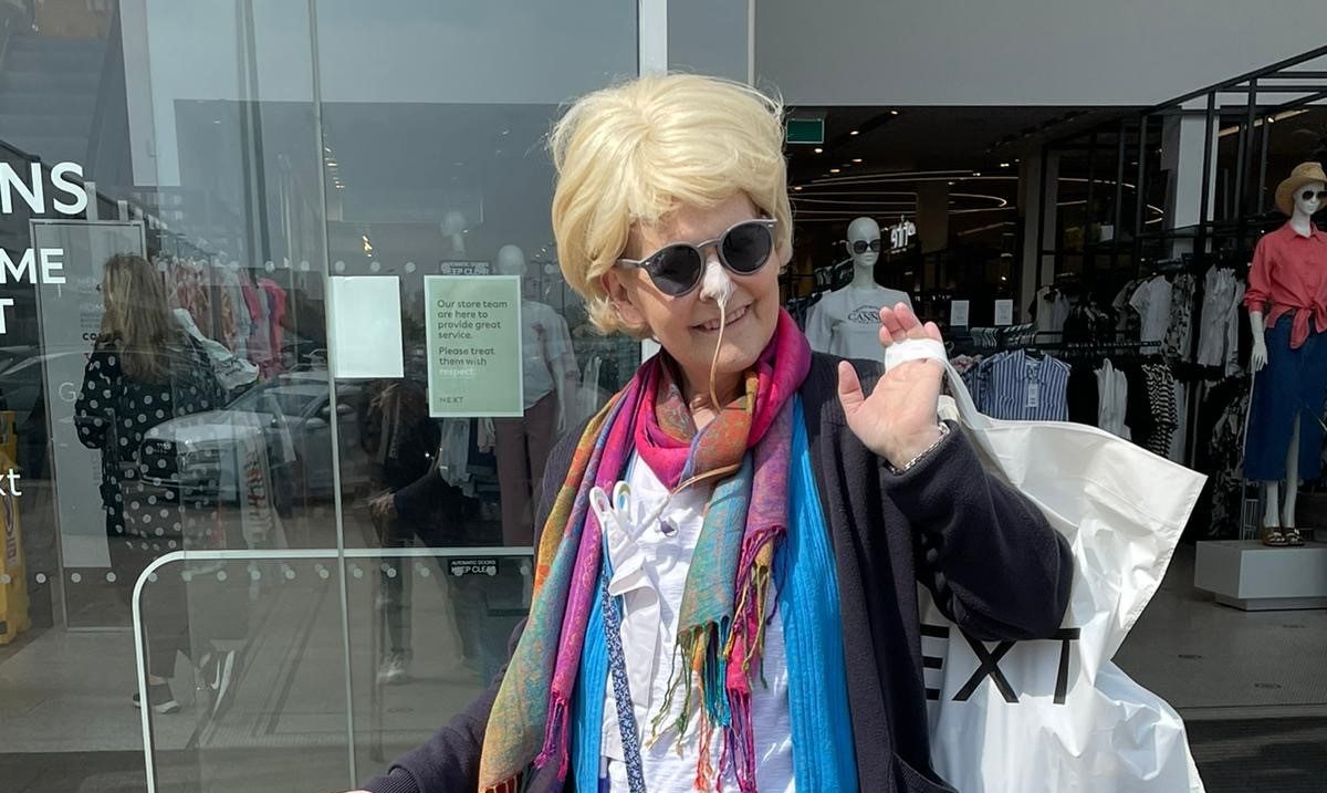 Susan stands outside a glass fronted shop with a shopping bag slung playfully over her shoulder. She is dressed in colourful clothes and is smiling at the camera.