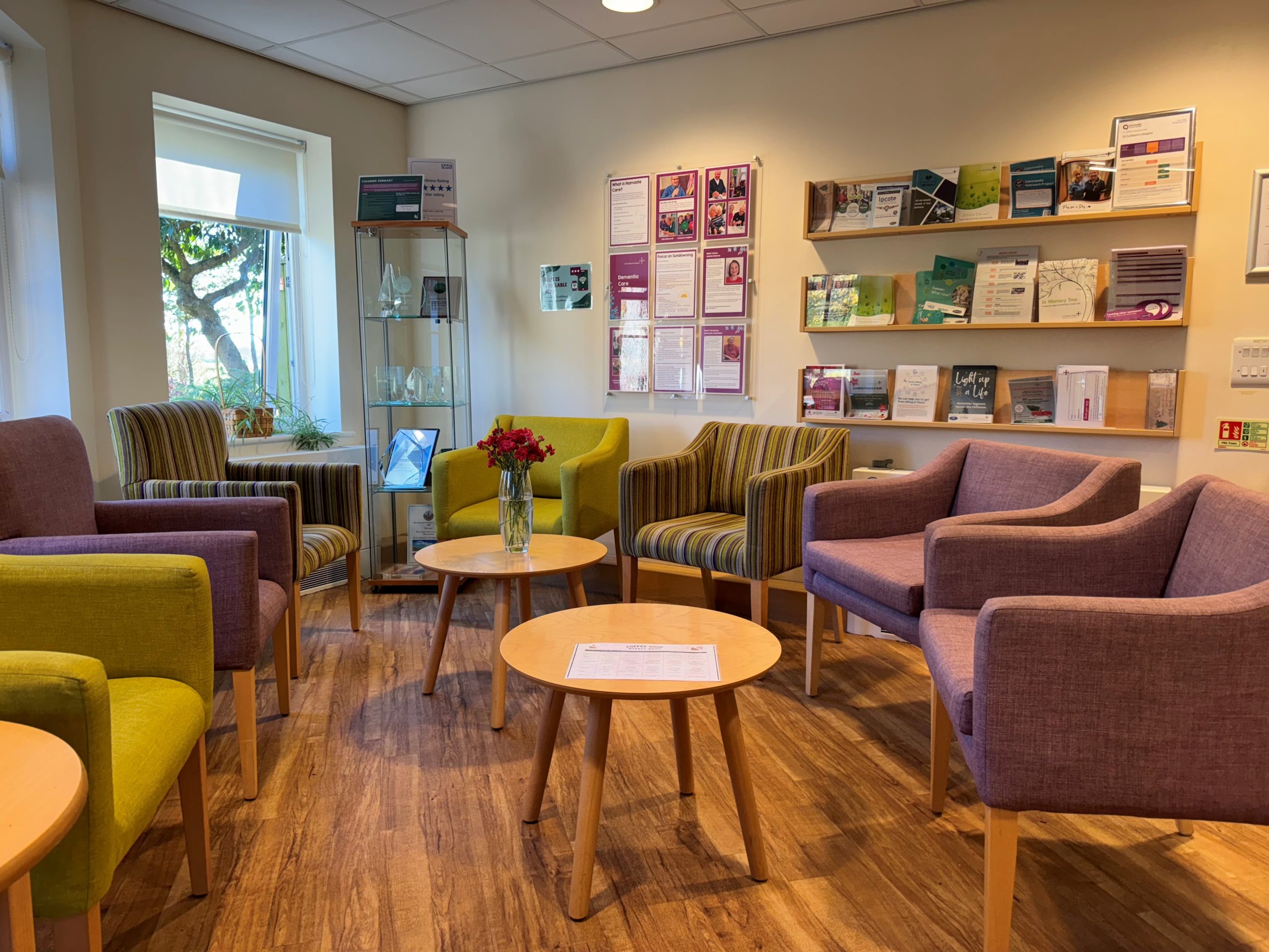 A cosy looking corner of a reception area with lots of brightly coloured seating and tables. Leaflet displays and windows in the background.