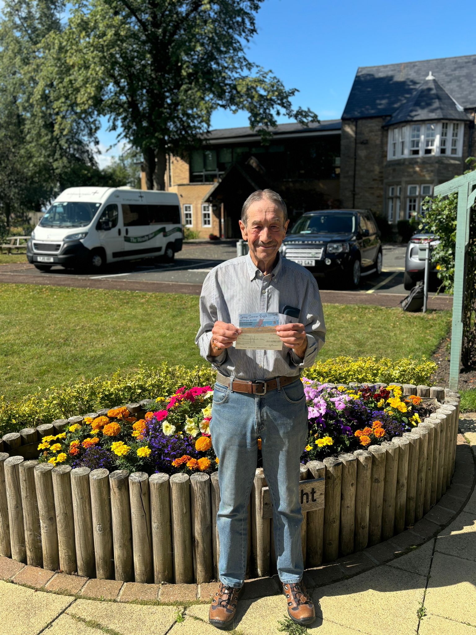 1. An older man smiles while holding a check in front of a colorful flower bed.