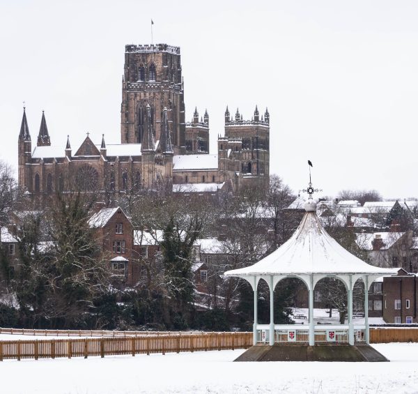 A bandstand surrounded by snow, with a Durham Cathedral rising in the background, showcasing a picturesque winter scene.
