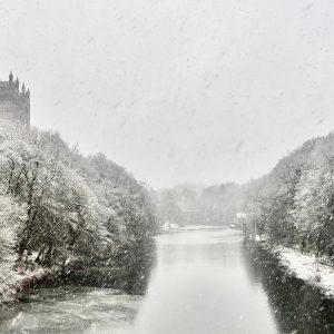A snowy river scene featuring trees and Durham Cathedral along the banks, creating a serene winter landscape.