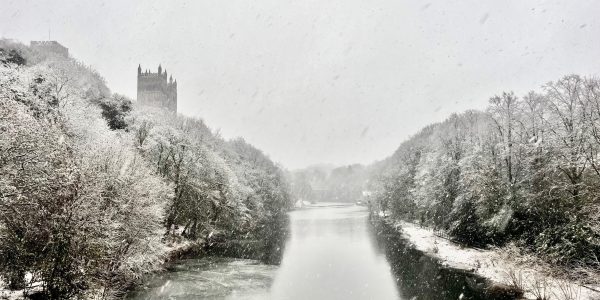 A snowy river scene featuring trees and Durham Cathedral along the banks, creating a serene winter landscape.