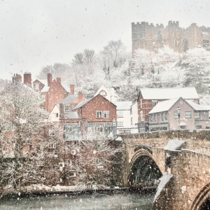 A snow-covered bridge spans a river, with gentle snowflakes falling around it, creating a serene winter scene.