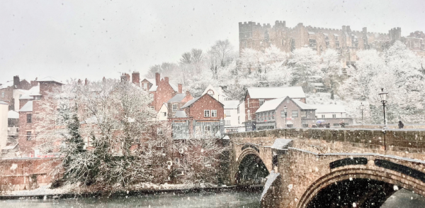 A snow-covered bridge spans a river, with gentle snowflakes falling around it, creating a serene winter scene.