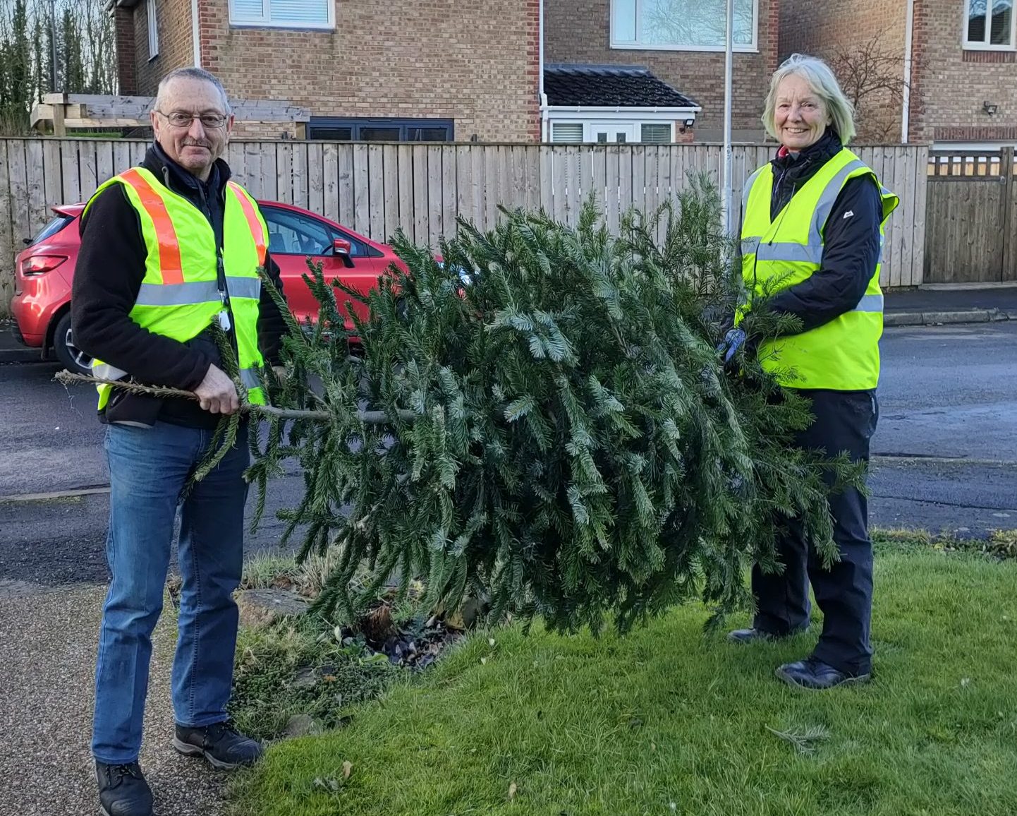 Two people wearing high vis vests stand holding a Christmas tree in a housing estate
