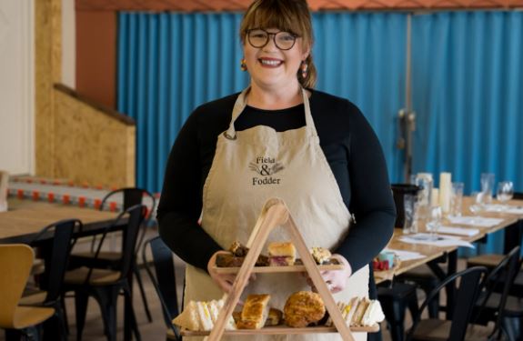A woman wearing glasses and an apron stands indoors, smiling and holding a wooden serving tray with assorted pastries and sandwiches.